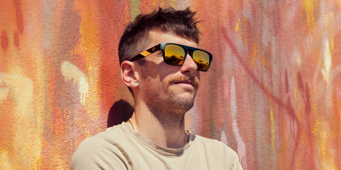 A man with short brown hair and a mustache stands against a vivid orange-pink graffiti wall, wearing Neven Eyewear’s Golden Banana sunglasses with polarized UV400 lenses. Relaxed in a beige t-shirt, he enjoys the sunlight, arms crossed.