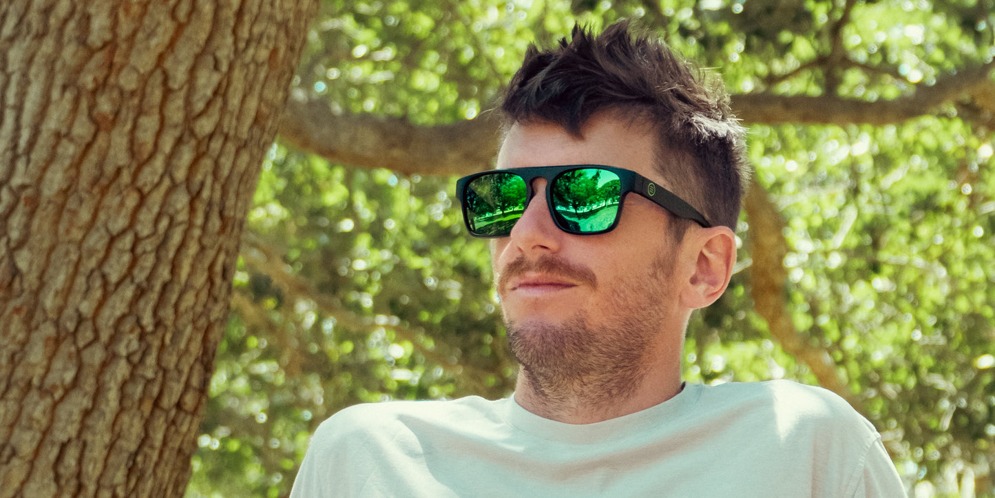 A man with short brown hair and stubble wears Neven Eyewear's Primatology black-framed polarized sunglasses with green mirrored lenses and a white t-shirt, leaning against a tree as sunlight filters through green leaves in the background.