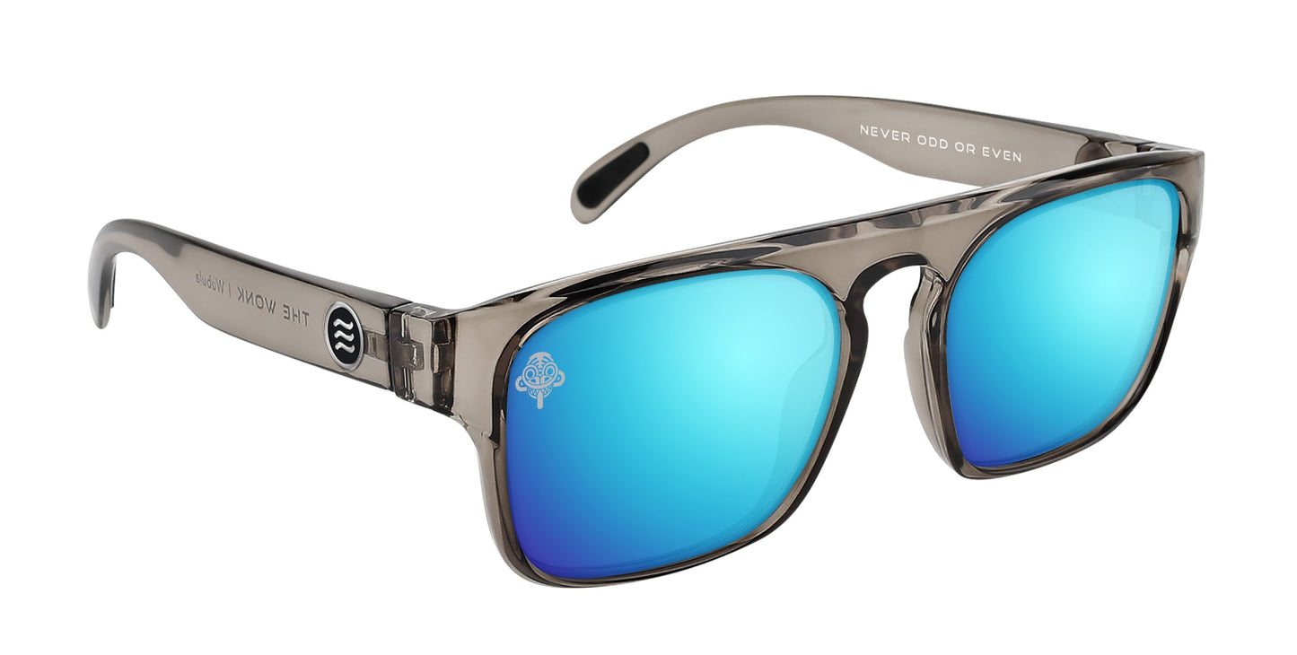 The Neven Eyewear Wubula sunglasses feature transparent gray frames, blue mirrored polarized lenses, a white emblem on the left lens, "NEVER ODD OR EVEN" inside the right arm, and a round Dirt Monkey logo on the outer left arm.