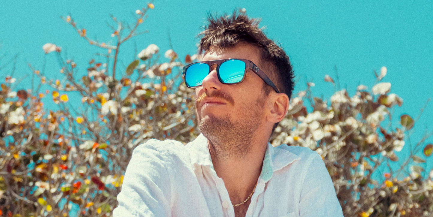 A man wearing a white shirt and Neven Eyewear's Wubula polarized sunglasses gazes upward with a relaxed smile. Sunlight highlights his face as leafy branches and a clear blue sky form a bright, summery backdrop.