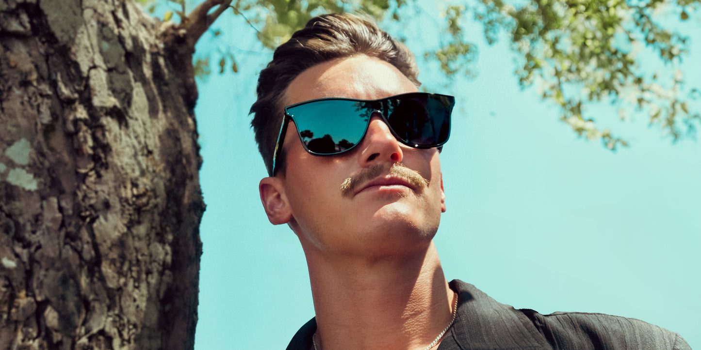 A man with short brown hair and a mustache wears Neven Eyewear's Goblin black polarized sunglasses with UV400 protection, standing by a tree in a dark shirt. Blue sky and green leaves form the background as he looks upward.