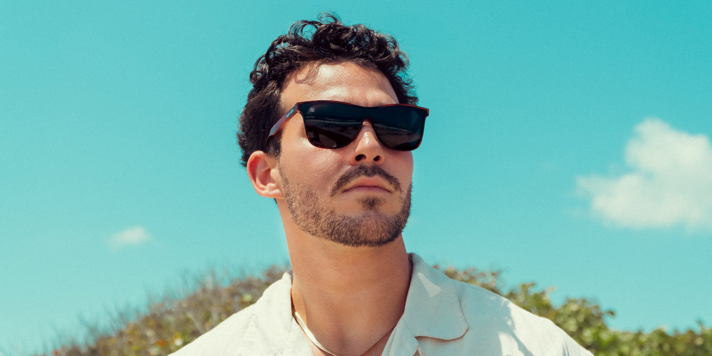 A man with short dark curly hair and a trimmed beard wears Neven Eyewear Warlock sunglasses with polarized UV400 protection and a light shirt, standing outdoors beneath a bright blue sky with green foliage in the background.