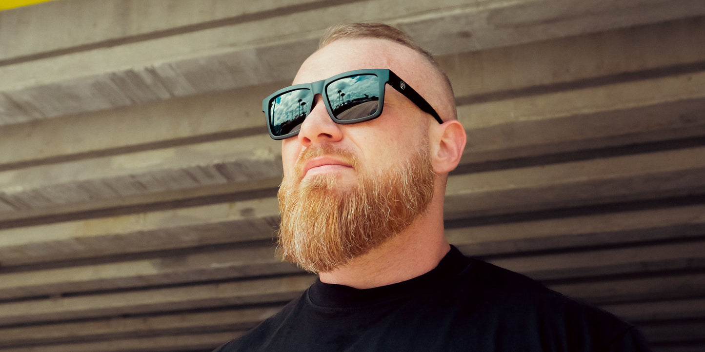 A light-skinned person with a reddish-blonde beard wears Neven Eyewear's Lowrider XL oversized rectangular sunglasses and a black shirt outdoors. Cloudy sky and railing reflections show in the lenses, with concrete beams behind them.