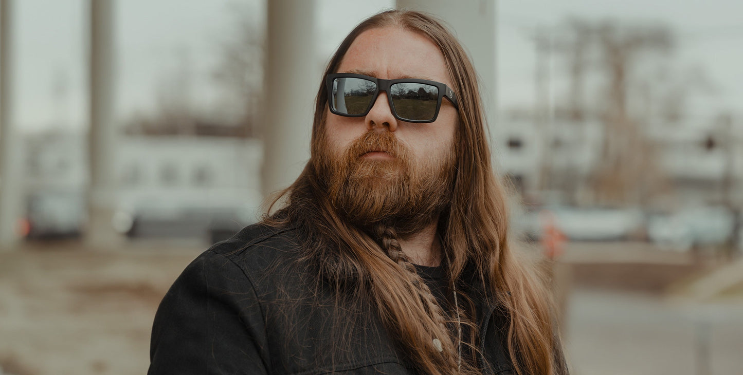 Wearing Neven Eyewear’s Ace sunglasses, a man with long brown hair and a full beard stands outdoors in a black jacket, with blurred buildings and trees behind him under an overcast sky.