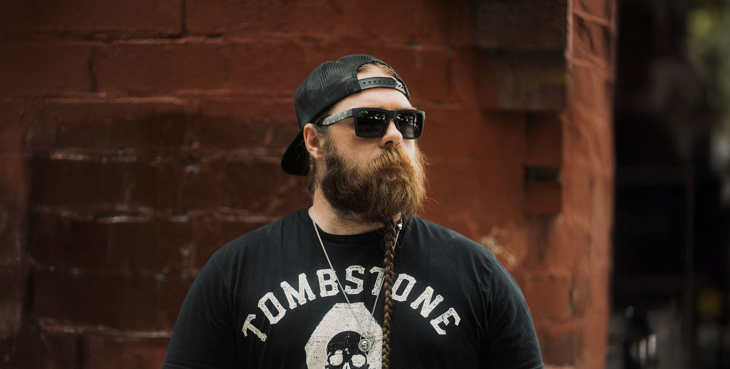 A bearded man with a long, side-braided beard wears Neven Eyewear’s Bad Company polarized UV400 sunglasses, a black backward cap, and a black Tombstone tee as he stands before a weathered red brick wall looking slightly right.