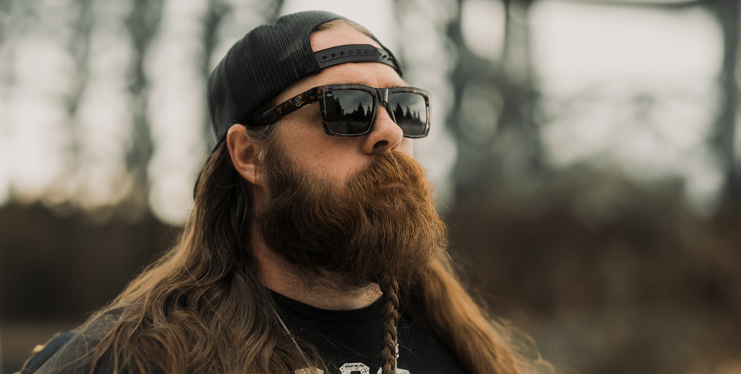 A man with long brown hair, a full thick beard, and a mustache wears a black baseball cap backwards, Neven Eyewear Jaguar sunglasses, and a black T-shirt. He stands outside with trees and an overcast sky behind him.