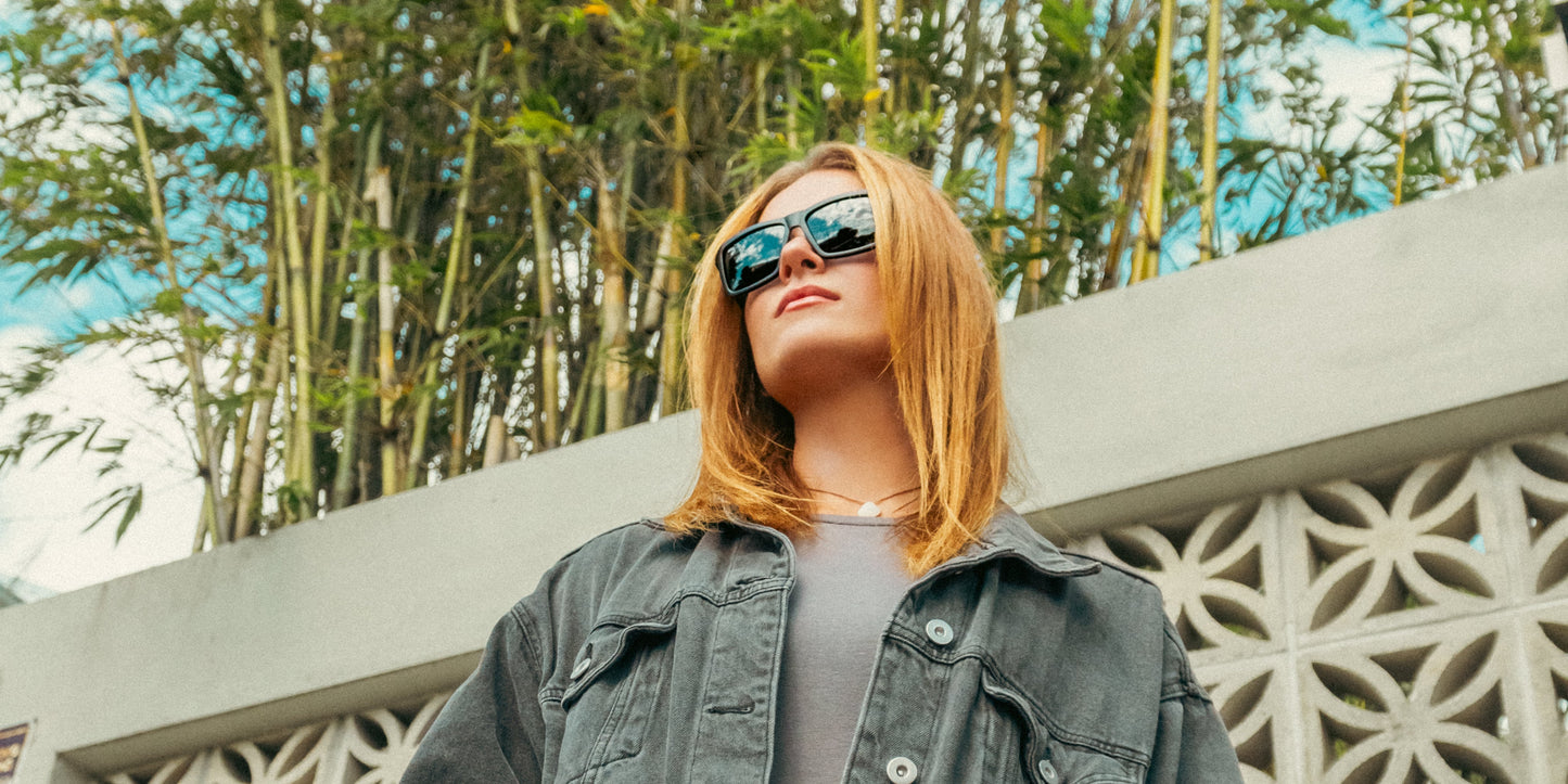 A person with shoulder-length blonde hair wears Neven Eyewear's Lowrider vintage motorcycle sunglasses, a gray shirt, and a dark denim jacket, standing outdoors in front of a white patterned wall and bamboo. Shot from below.