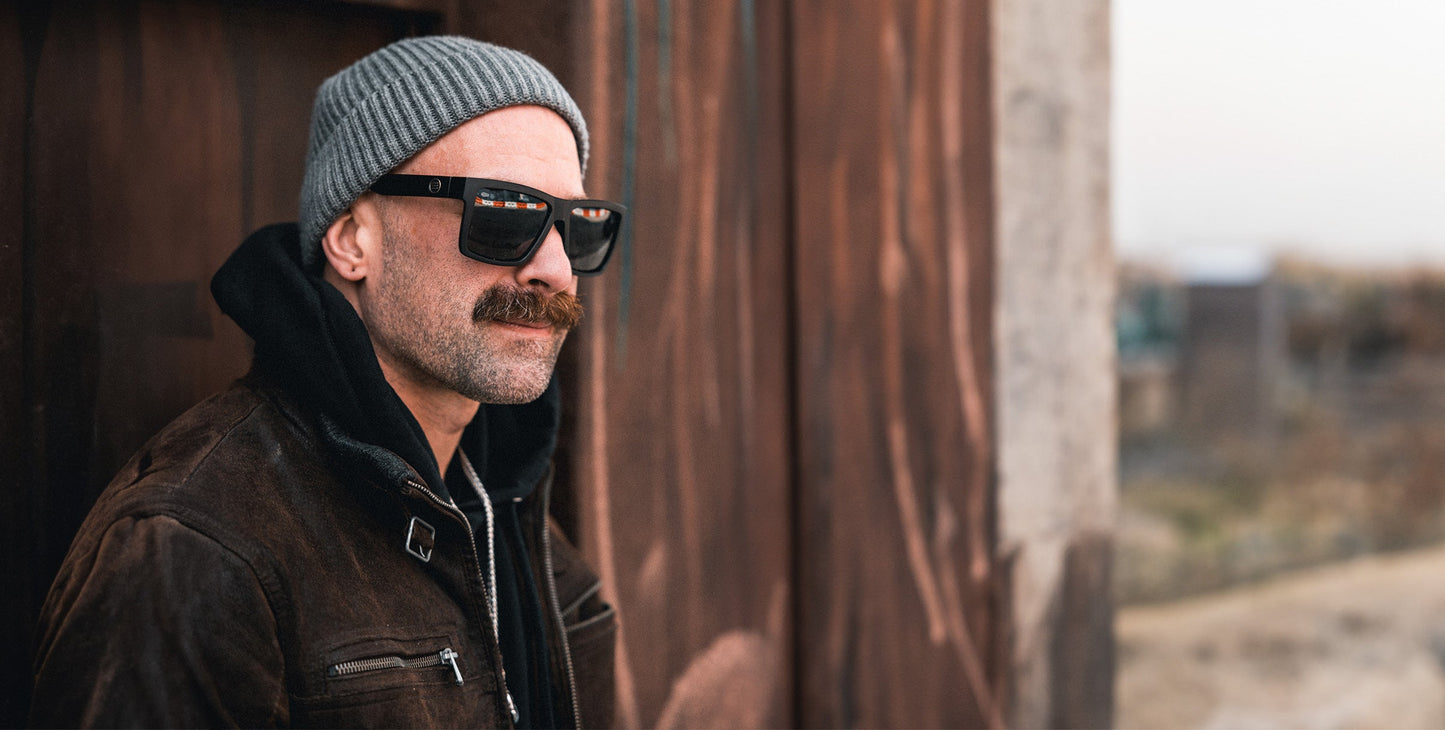A man with a thick mustache wears a gray beanie, a dark brown jacket with a hoodie, and Neven Eyewear’s Lowrider vintage motorcycle sunglasses as he stands outside against a rust-colored metal wall.