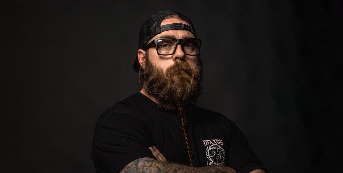 A bearded man with a long braid stands arms crossed, wearing a black t-shirt, backward black cap, and Neven Eyewear Malibu clear motorcycle glasses. Colorful tattoos cover his forearms as the dark background highlights his serious expression.