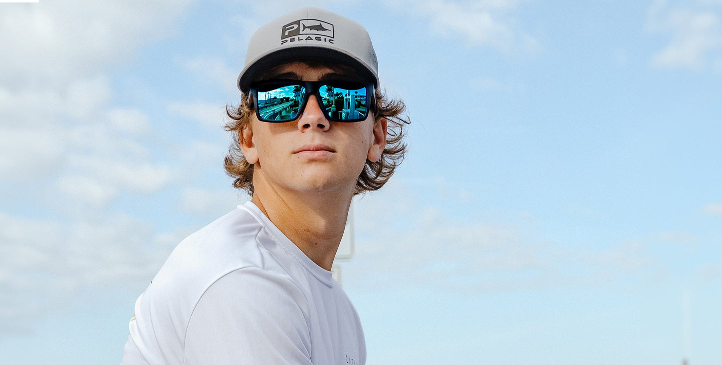 A young man outdoors looks to the side, wearing a light gray cap and Neven Eyewear's Montauk glasses with black frames. His reflective sunglasses mirror a boat scene, and he has light brown wavy hair. The sky is blue with scattered clouds.