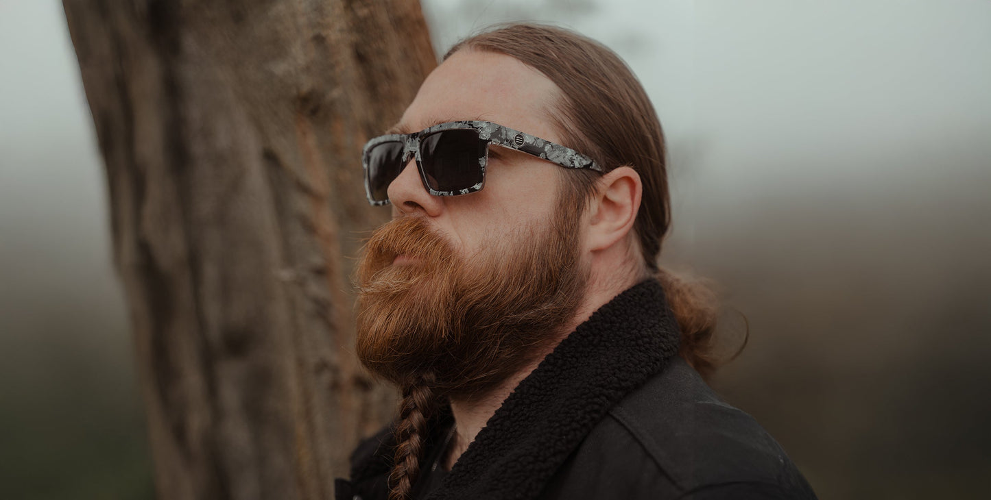 A bearded person with long, reddish-brown braided hair stands near a tree outdoors, wearing Neven Eyewear’s South Shore sunglasses and a black jacket with a fuzzy collar. The blurred, misty background adds a soft atmosphere.