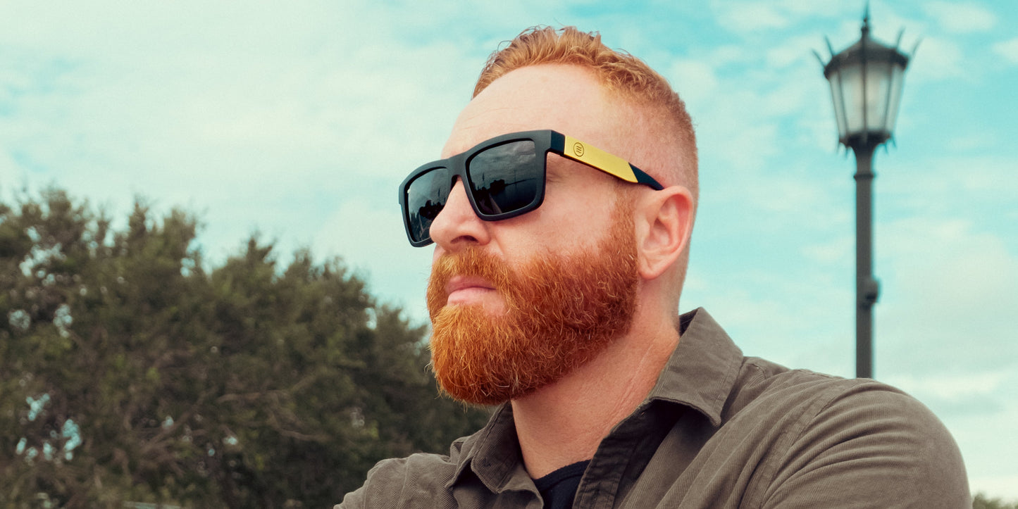 A man with a full red beard and short red hair wears Neven Eyewear's Prospector XL sunglasses with polarized black lenses and a gray collared shirt, gazing into the distance as a lamp post, trees, and partly cloudy sky complete the backdrop.
