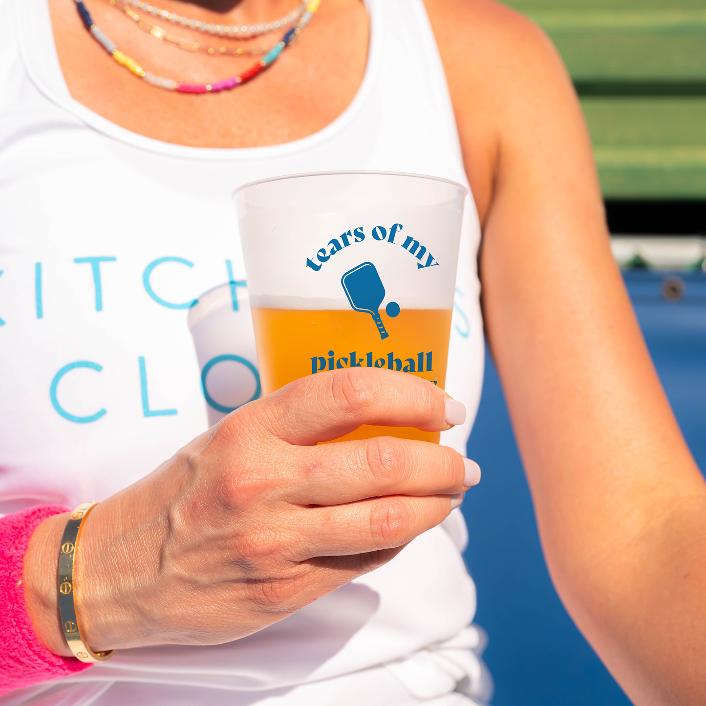 A woman wearing a white tank top and colorful beaded necklace holds a Swinton Pickleball "Tears of my Pickleball Opponents" reusable party cup filled with an orange drink next to a blue paddle graphic, sporting a pink wristband and gold bracelet.
