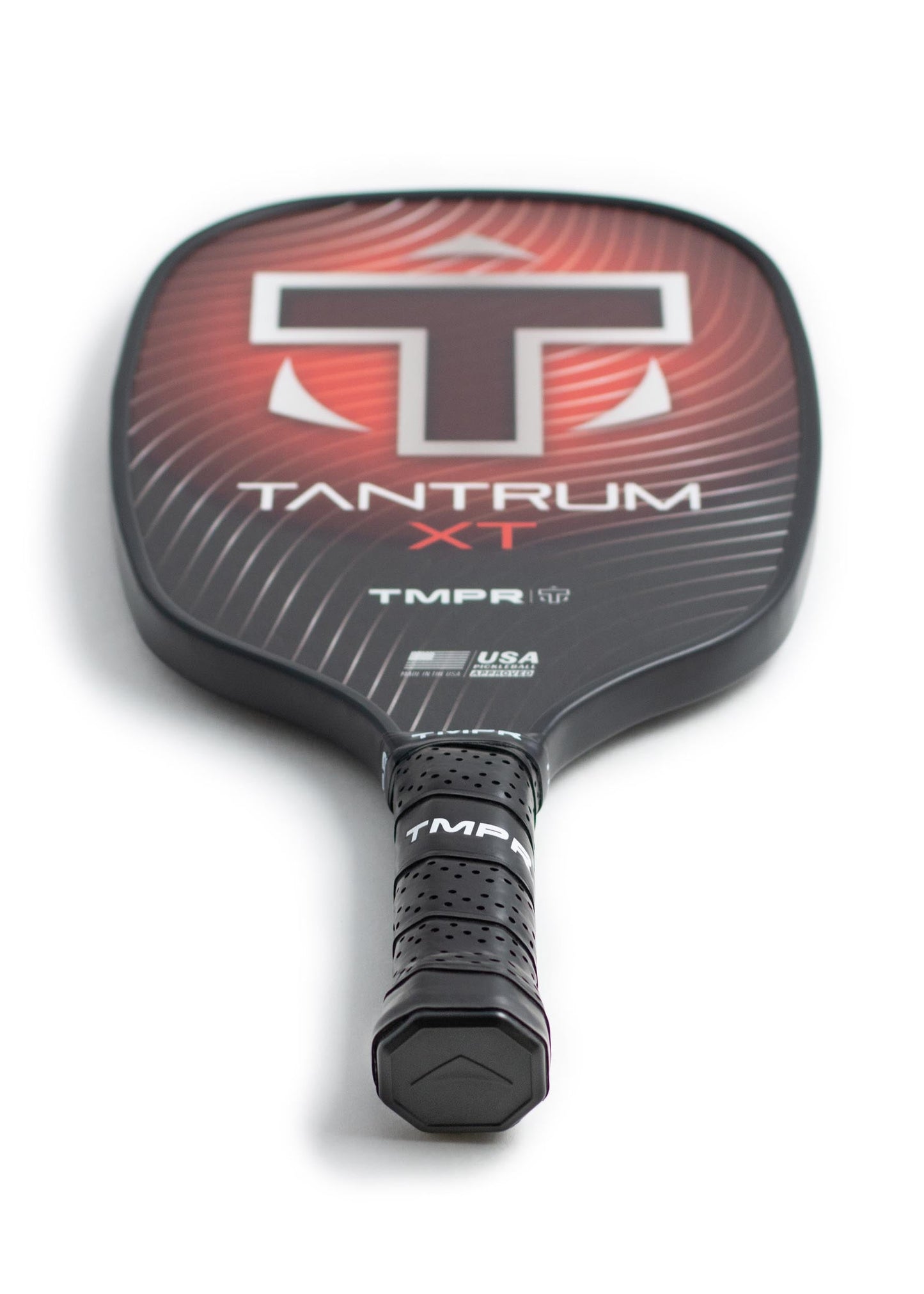 A close-up of the TMPR Sports Tantrum XT pickleball paddle shows its black handle, perforated grip, and striking red and black face with a large white T logo. The honeycomb polymer core delivers excellent performance. Handle is shown in a forward position.