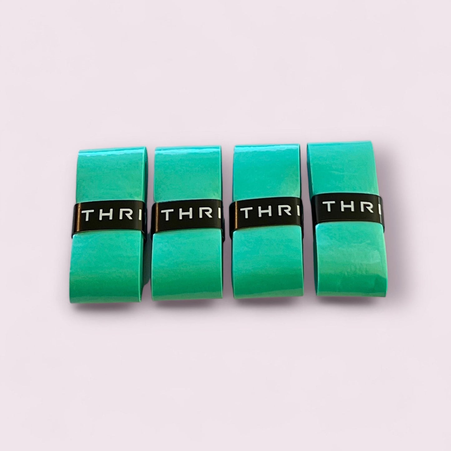 Four glossy green rectangular Overgrips (Super Tacky) from Thrive Pickleball are arranged in a row on a pale pink surface, each with a black band featuring white "THRI" letters, highlighting their tacky and durable design for peak performance.