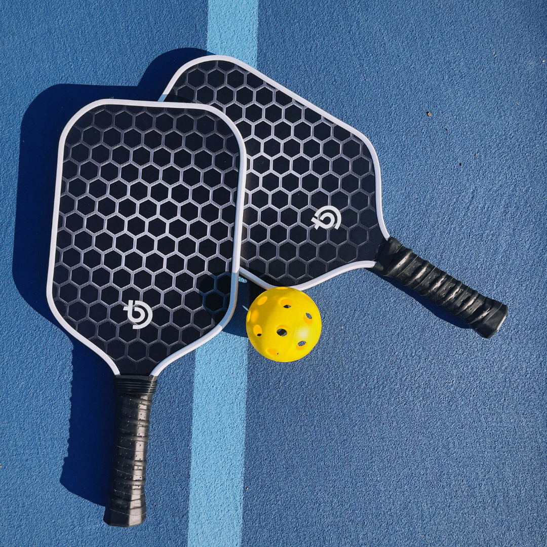 The Big Dink Bundle by The Big Dink features two black carbon fiber pickleball paddles with honeycomb patterns overlapping on a blue court, handles outward, and a yellow ball between them near a vertical light blue dividing line.