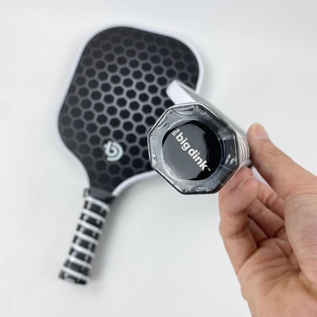 A hand holds The Big Dink paddle by the handle, showing its branded end cap. The paddle, part of The Big Dink Bundle, has a black honeycomb pattern with white trim; another similar one sits on a white surface in the background.