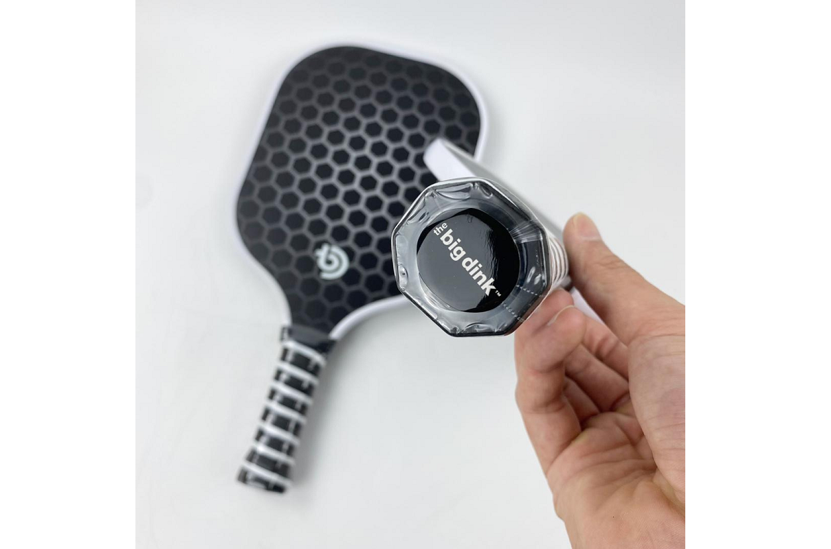 A hand grips The Big Dink - Model1 Paddle, featuring a black hexagonal honeycomb pattern. The handle’s end cap shows The Big Dink logo in white. Two paddles are included in this 2-pack special, displayed on a white background.