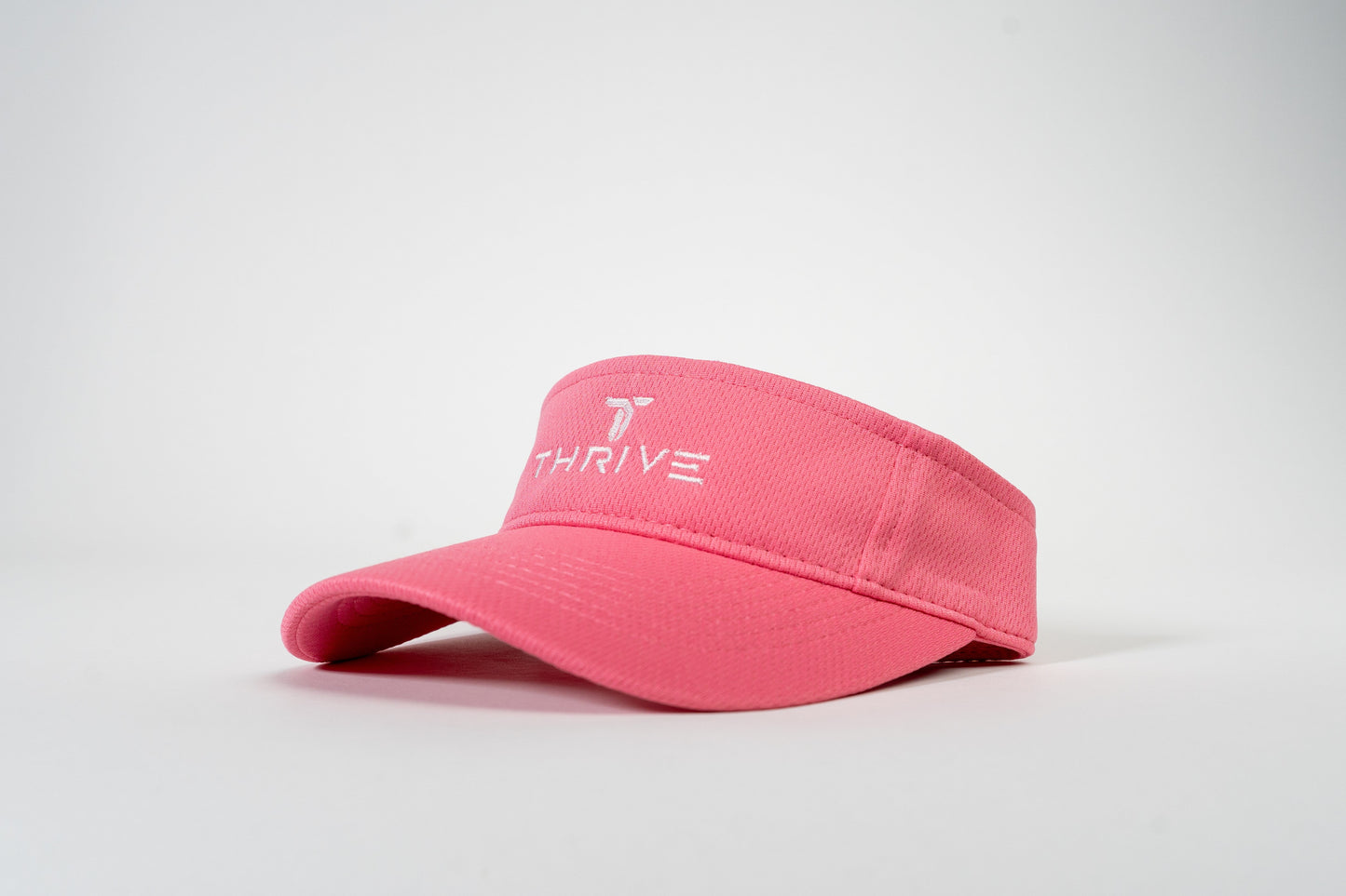The Thrive Elite Tour Visor by Thrive Pickleball is a pink, moisture-wicking visor featuring "THRIVE" and a white, stylized T logo embroidered on the front panel. It's shown against a white background, angled to highlight its texture and stitching.