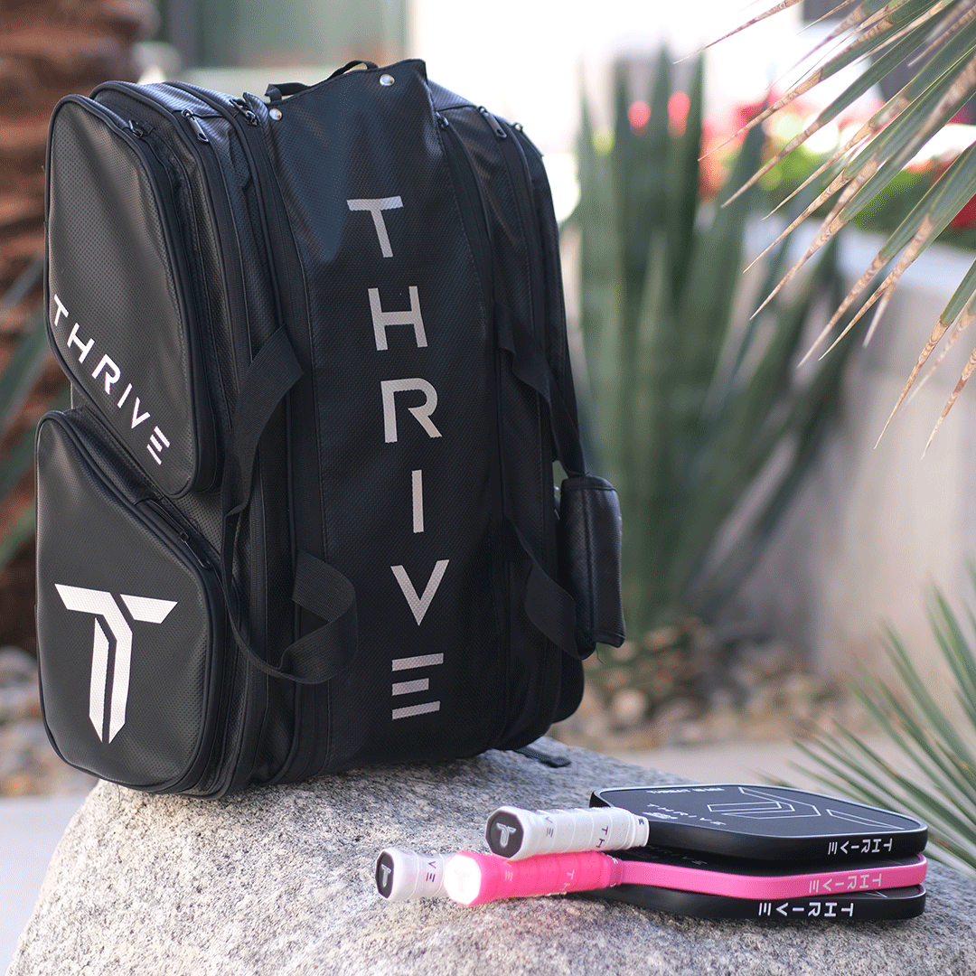 A black Elite Pro Tour Bag w/shoe bag by Thrive Pickleball, featuring white logos, sits outdoors with two paddles and balls in front—one paddle has a pink grip. Perfect for stylishly carrying your gear to the courts.