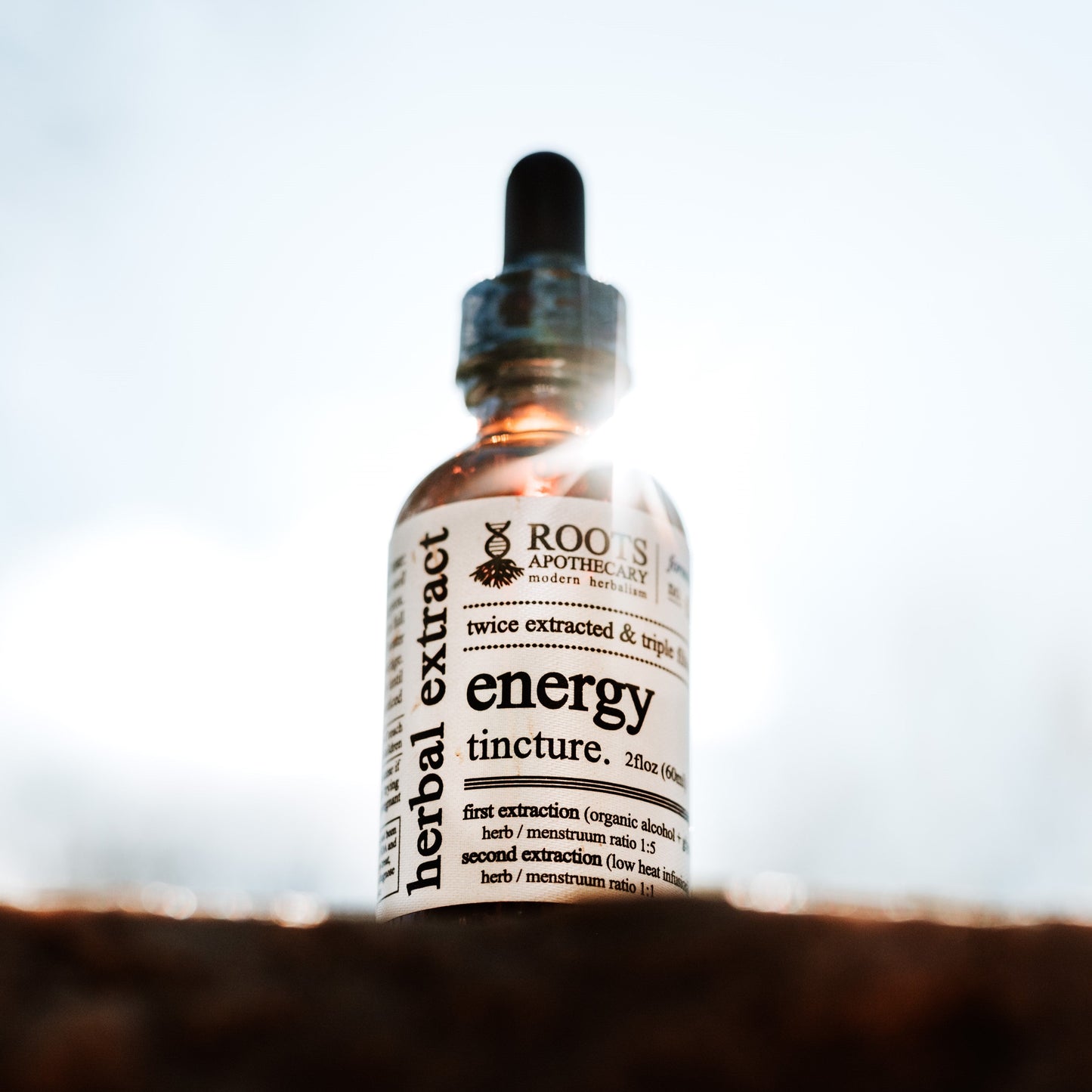 A Roots Apothecary energy tincture glass dropper bottle sits on a wooden surface, sunlight highlighting its label featuring herbal extracts for memory and focus support, including ashwagandha, rhodiola, and organic green tea.