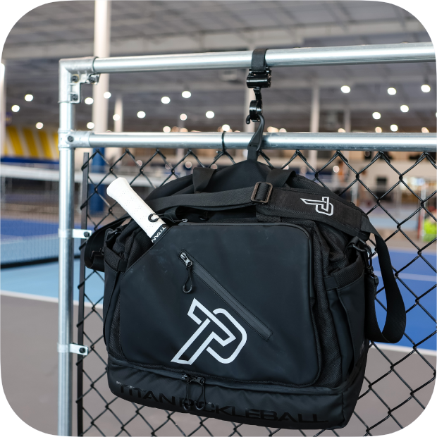 The Titan Pickleball Titan Duffle Bag in black, featuring a white logo, hangs on a chain-link fence. A white paddle is tucked in the side pocket, with blue indoor courts and a sports facility visible in the background.