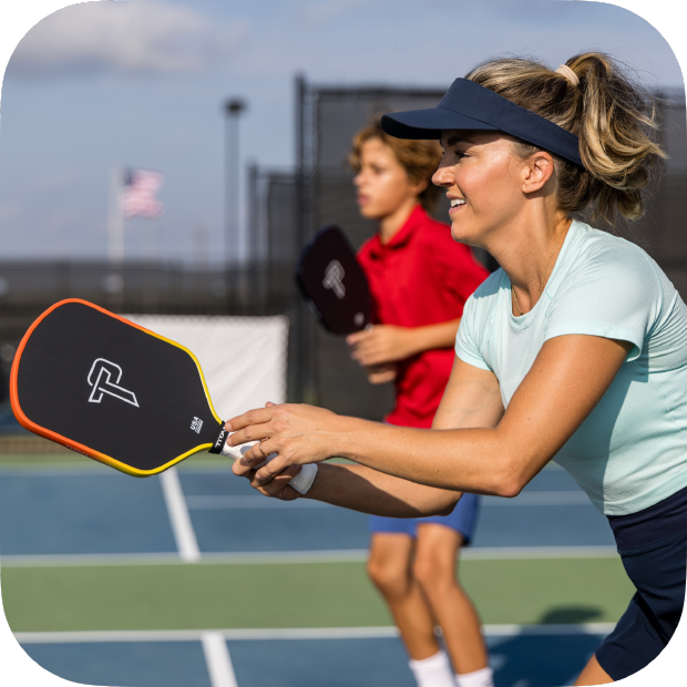 A woman in a light blue shirt plays outdoor pickleball with the Titan Ignite 16L paddle by Titan Pickleball, smiling as she leans forward. A boy in a red shirt and blue shorts stands behind her on a blue and green court, with an American flag in the background.