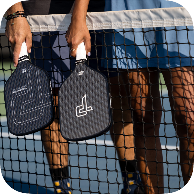 Two people stand side by side on a blue and white pickleball court near the net, holding Titan Pickleball's Titan Pro Titanium 14L paddles with white geometric designs. Only their lower bodies and paddle-holding hands are visible.