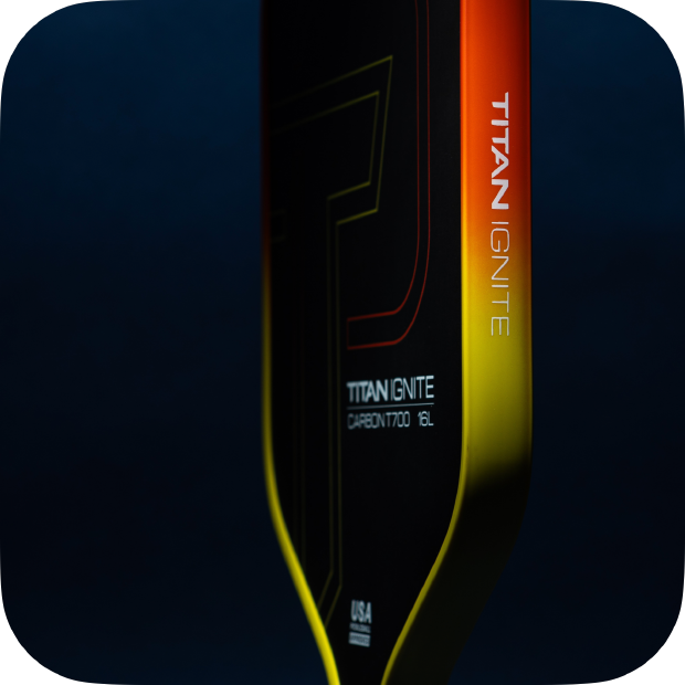 Close-up of the Titan Ignite 16L paddle by Titan Pickleball, featuring an elongated carbon fiber face with a yellow-to-orange gradient, visible TITAN IGNITE and CARBON F700 EL text, faint T outline, USA emblem, and a dark blue background.