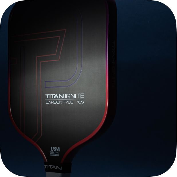 A close-up of the Titan Pickleball Titan Ignite 16S carbon fiber pickleball paddle with red and purple accents. Text on the paddle: "TITAN IGNITE CARBON T700 16S" and "USA PICKLEBALL APPROVED." Deep blue background. Ideal for buyers.