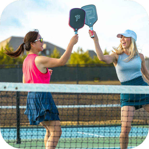 Two women celebrate on an outdoor pickleball court, tapping their Titan Pickleball Titan Ignite 16S carbon fiber paddles above the net. One wears a pink top and navy skirt, the other a blue tank, teal skirt, and white cap. Both are smiling.
