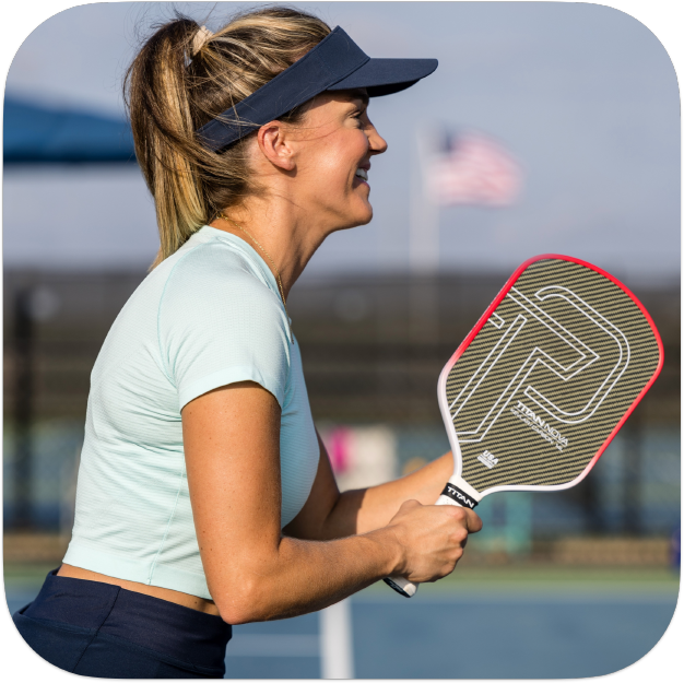 A woman in a light blue shirt and visor smiles as she holds the Titan Pickleball Titan Nova 14L paddle, ready for action on an outdoor court with an American flag and net in the blurred background.