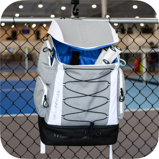 A gray and white Titan Pickleball Backpack from Titan Pickleball hangs on a fence by an indoor court. The open bag shows a blue interior with a white water bottle inside, and black bungee cords cross the front pocket.