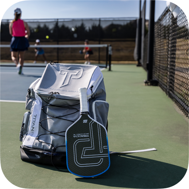 A Titan Pickleball Backpack rests on the court near a chain-link fence, alongside a black-and-white paddle, as two people play in the background. The spacious Titan Pickleball bag is ideal for carrying all your gear on sunny days.
