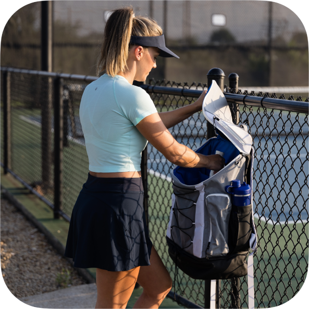 A woman in a light blue top, navy skirt, and visor stands by a tennis court fence, placing items into the Titan Pickleball Backpack from Titan Pickleball hanging nearby as sunlight casts shadows across her and the ground.