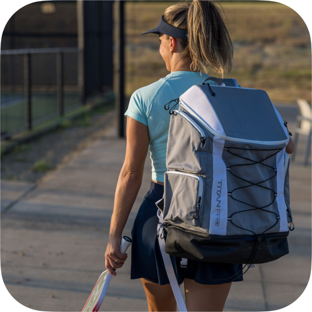 A woman with a ponytail walks on a sidewalk near a tennis court, holding a paddle and carrying the Titan Pickleball Backpack by Titan Pickleball. She wears a blue cap, light blue top, and navy skort, heading away from the camera in sunlight.