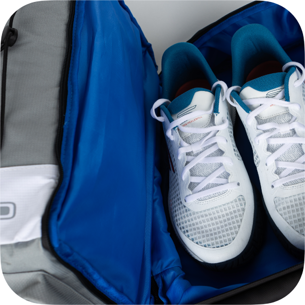 A pair of white athletic shoes with teal accents sits neatly inside the spacious, blue-lined compartment of a gray Titan Pickleball Backpack by Titan Pickleball, which is unzipped and open. The clean shoes and rounded image corner are visible.