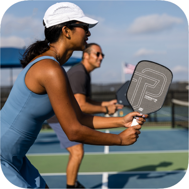 A woman in a blue athletic dress and white visor holds a Titan Pickleball Titan Pro Titanium 14L paddle, ready to play on an outdoor court, while a man in dark sportswear stands behind her. The sky is clear and the American flag waves in the background.