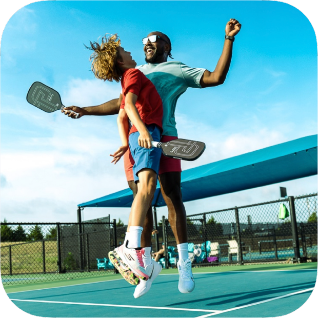 Two people celebrate on an outdoor pickleball court, jumping and bumping chests with Titan Pro Titanium 16L paddles by Titan Pickleball in hand. Both wear athletic gear and sneakers. Blue canopy, fence, and partly cloudy sky complete the scene. Shop now!.