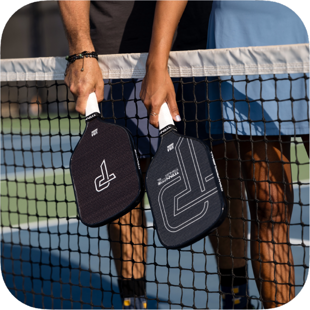 Two people stand on opposite sides of a tennis net, each holding a Titan Pickleball Titan Pro Titanium 16L paddle with white handles downward. The focus is on their hands and paddles; only arms and legs show against a blue and green court backdrop.