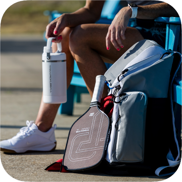 A person with red nail polish sits on a blue bench, wearing white sneakers and a smartwatch, holding a white water bottle. Next to them is a gray sports bag with the Titan Pro Titanium 16L pickleball paddle by Titan Pickleball partially visible.