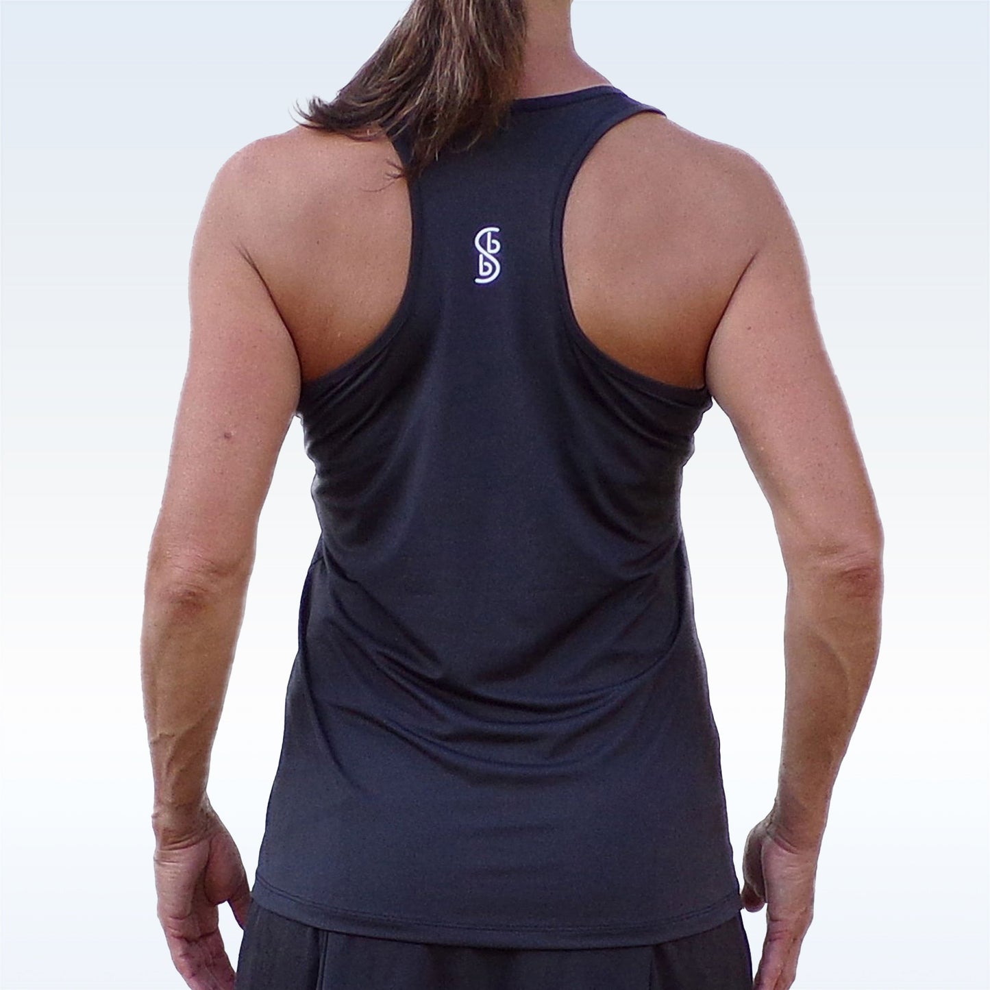 A person with long brown hair in a ponytail is shown from behind, wearing the Pickleball Bella PB Racerback Tank Black, which features a racerback design and small white logo on the upper back. The background fades from light blue to white.