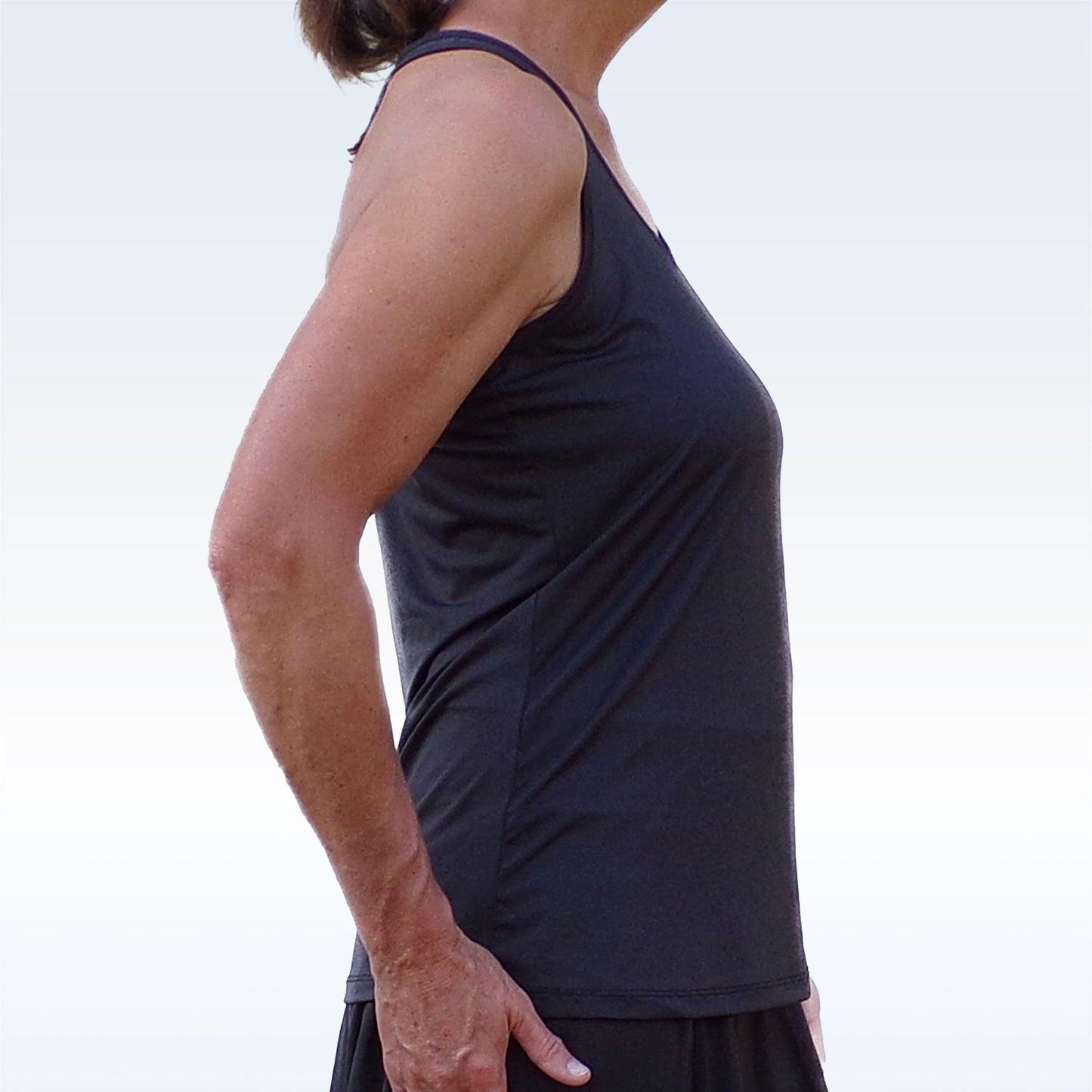 A woman stands in profile against a light background, wearing the Pickleball Bella PB Racerback Tank Black and a black skirt. Her left arm rests at her side, and her shoulder-length brown hair completes the look from shoulders to hips.