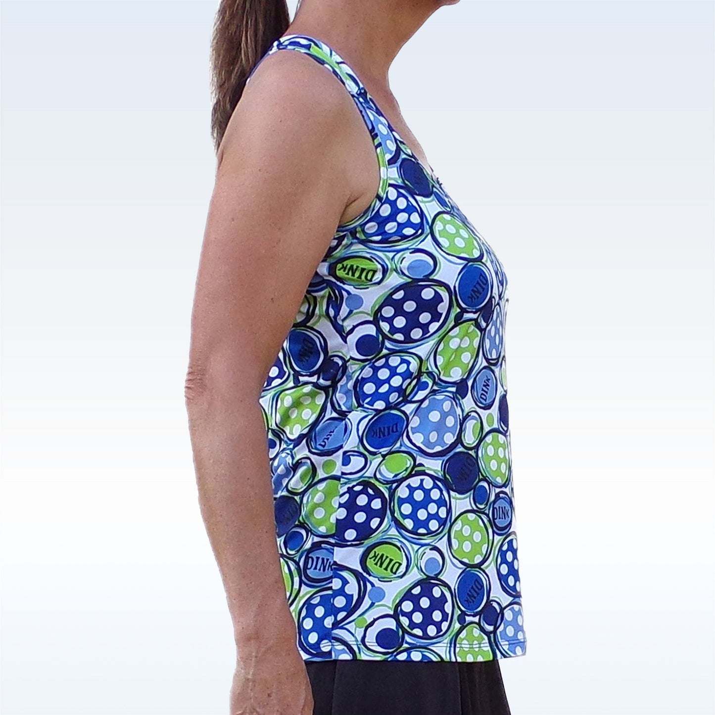 A woman stands in profile wearing Pickleball Bella’s PB Racerback Tank Dink 1, featuring a blue, green, and white tennis ball pattern with “DINK” print. Made from performance fabric, she pairs it with dark shorts against a light background.