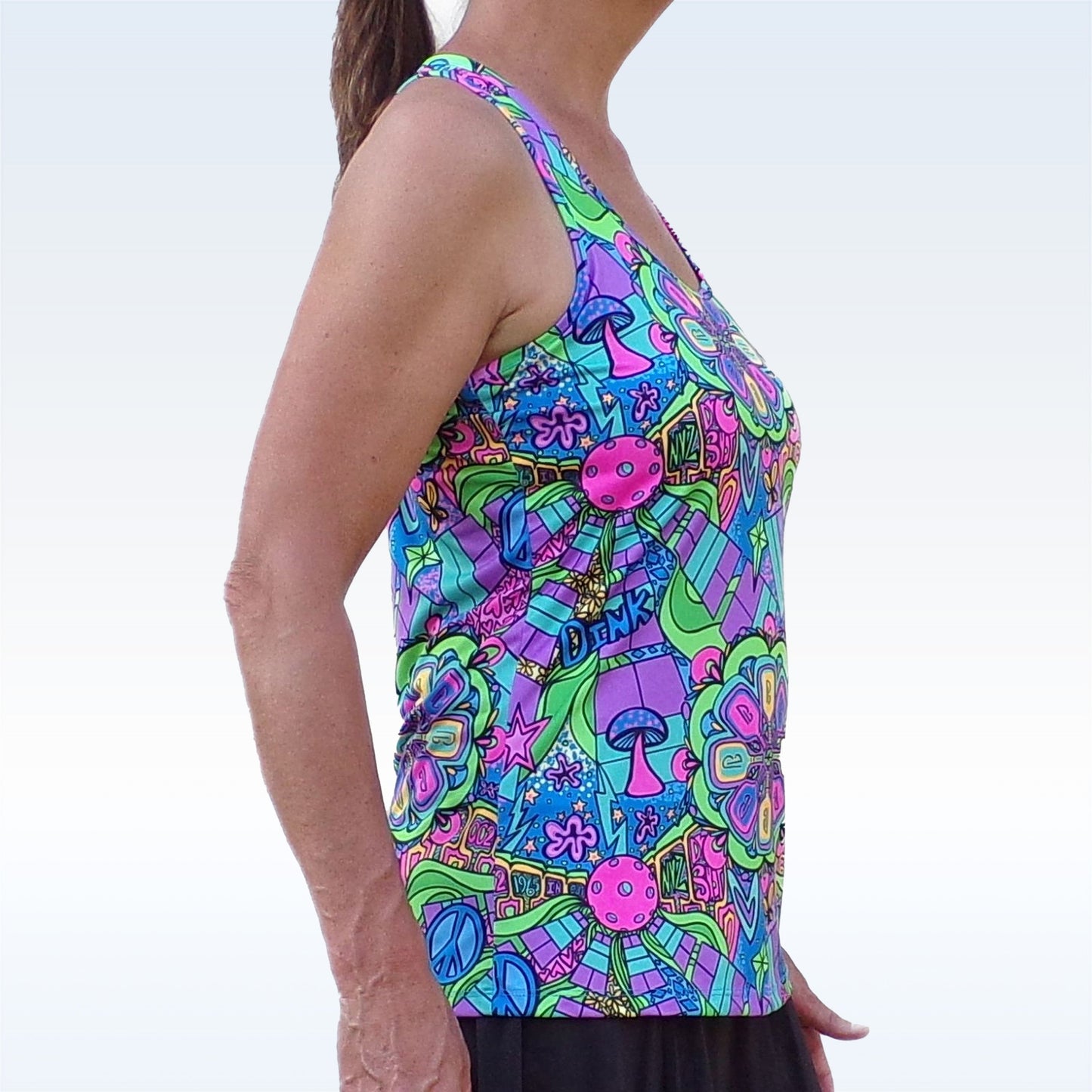 A woman stands in profile against a light blue background, wearing the Pickleball Bella PB Racerback Tank Groovy. Its vibrant psychedelic pattern with flowers, mushrooms, stars, and peace signs offers style and comfort in performance fabric for pickleball.