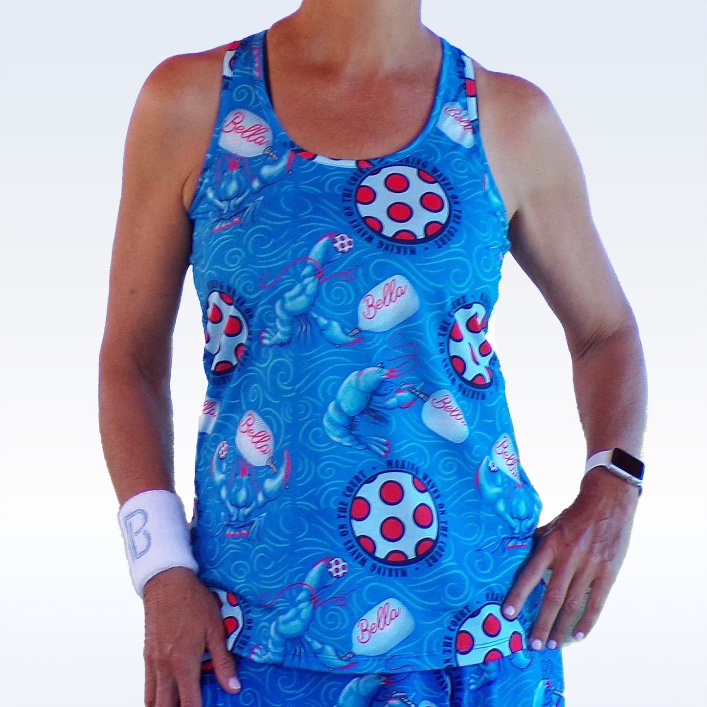 A person wears the Pickleball Bella PB Racerback Tank Lobster Monster, featuring blue fabric with red and white polka dot targets, lobsters, and "Bella" print. They also sport a white B wristband and a smartwatch against a light gradient background.