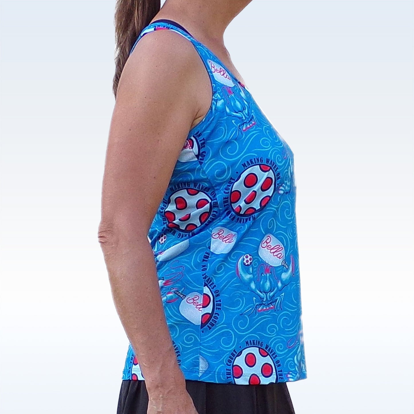 A woman with light skin and a ponytail wears the Pickleball Bella PB Racerback Tank Lobster Monster, a sleeveless blue top with playful patterns, paired with a black skirt. She's shown from the side against a pale blue background.