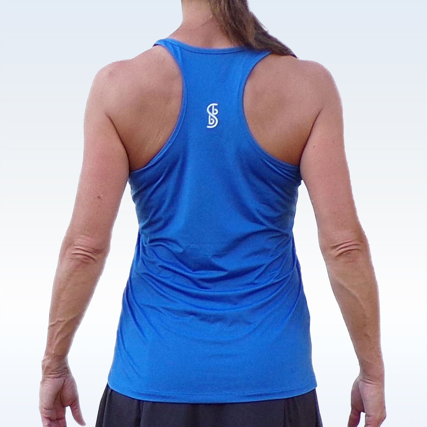A person with long hair in a ponytail is shown from behind wearing the Pickleball Bella PB Racerback Tank in Royal Blue, featuring a racerback design and white logo between the shoulders, set against a light gradient background.