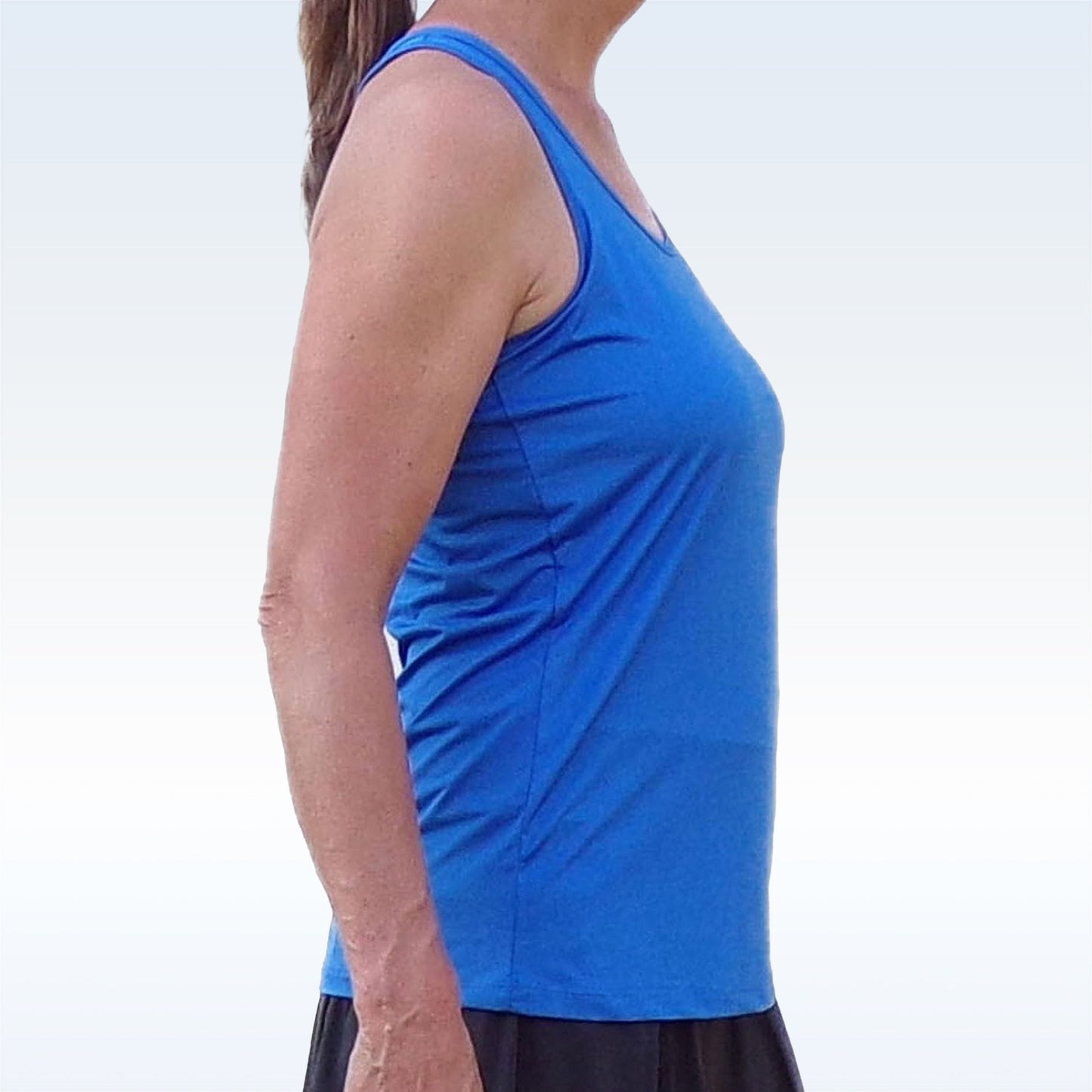 A person in profile faces left, wearing the Pickleball Bella PB Racerback Tank in Royal Blue and black shorts. Their long brown hair is tied back in a ponytail. The background is plain and light, with no visible objects.