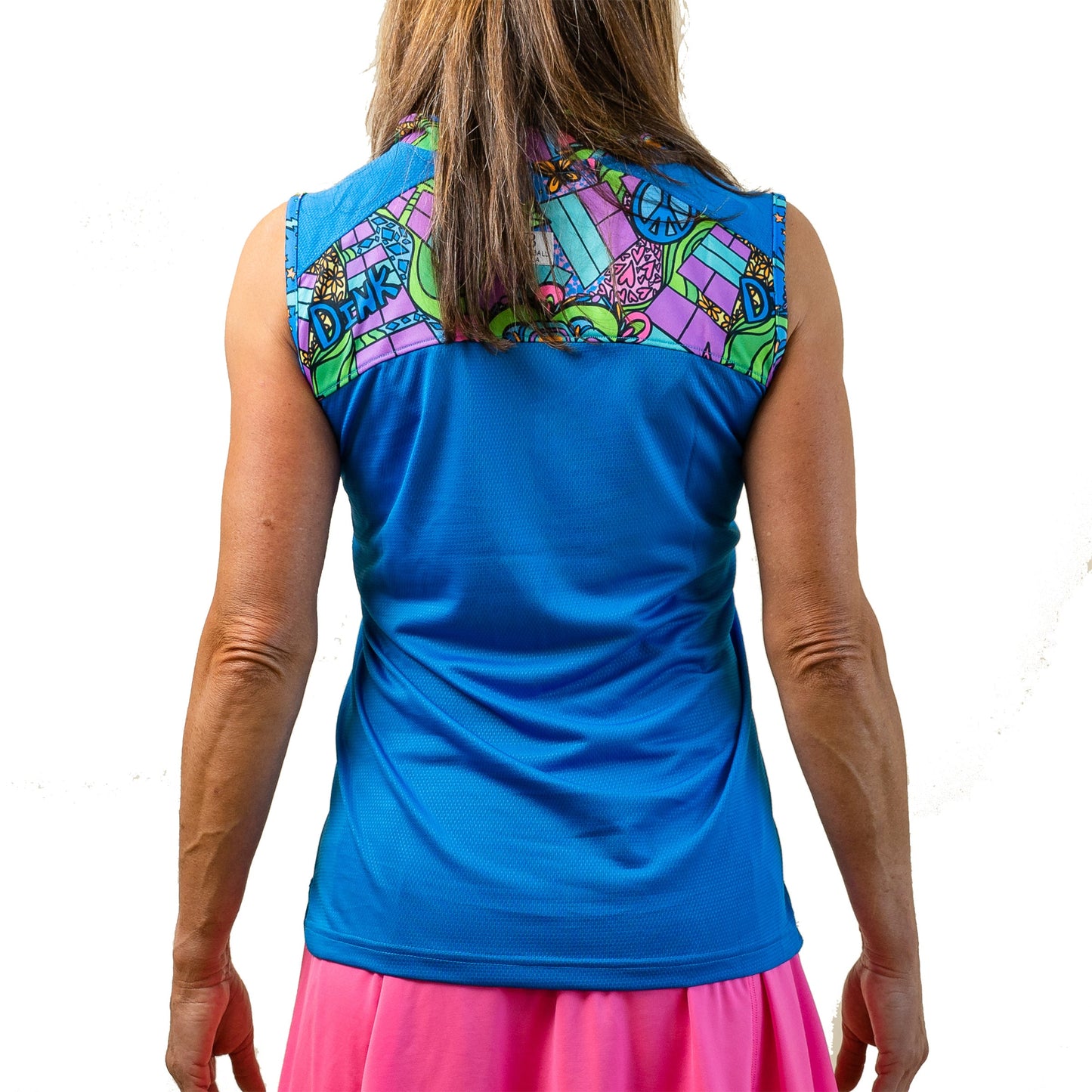 A woman stands facing away on a white background, wearing the Pickleball Bella PB 1/4 Zip Sleeveless Groovy top with a colorful, patterned yoke of peace signs and hearts, plus a pink skirt. Her brown shoulder-length hair and arms are visible.